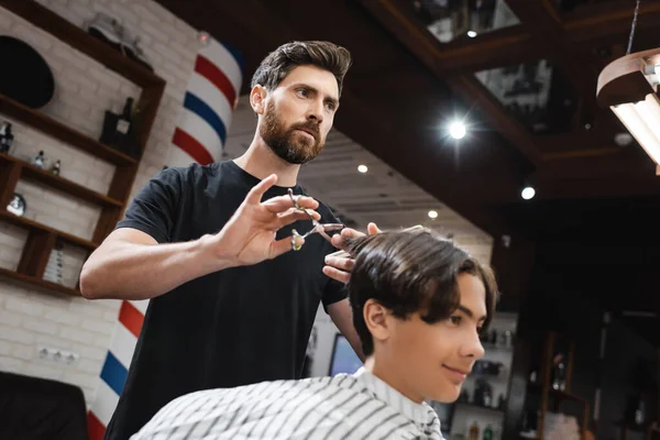 Bajo ángulo vista de barbero barbudo haciendo corte de pelo a cliente adolescente en salón de belleza - foto de stock