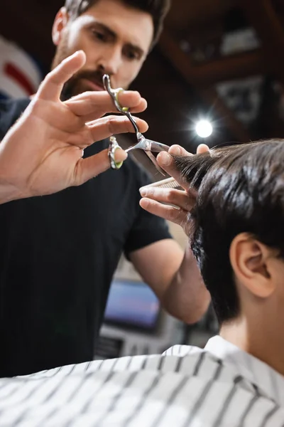 Low angle view of blurred hairdresser cutting hair of teenage client in barbershop — Stock Photo