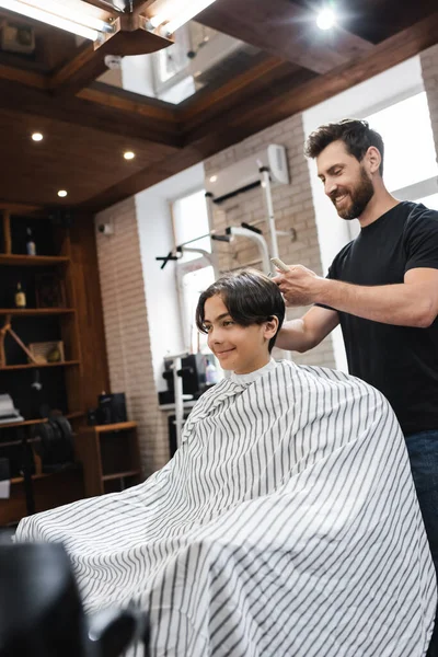 Парикмахер с бородой работает с клиентом-подростком, сидящим в парикмахерской в салоне красоты — стоковое фото