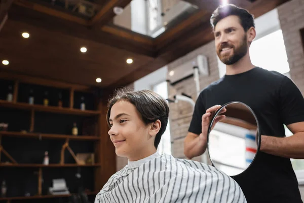 Feliz cabeleireiro segurando espelho perto de cliente adolescente satisfeito na barbearia — Fotografia de Stock