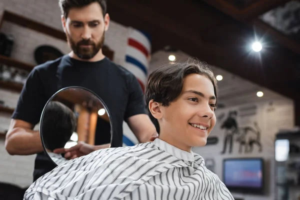 Cabeleireiro desfocado segurando espelho perto de menino adolescente feliz sentado na capa de cabeleireiro — Fotografia de Stock