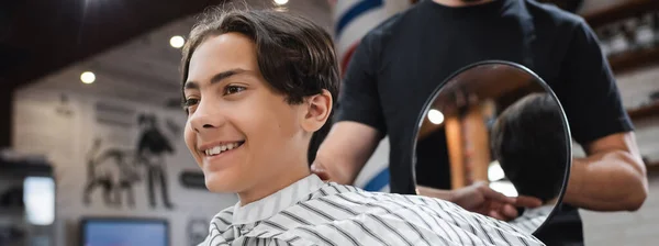 Alegre adolescente sonriendo cerca de peluquería con espejo en la barbería, pancarta - foto de stock