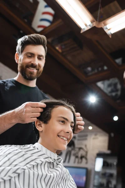 Low angle view of smiling barber touching hair of happy brunette teenager in beauty salon — Stock Photo