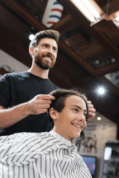 Vista de ángulo bajo de adolescente alegre cerca de peluquero tocándose el pelo en la barbería - foto de stock