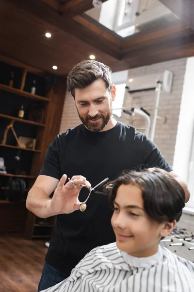 Cabeleireiro barbudo positivo trabalhando com cliente adolescente sorrindo em primeiro plano desfocado — Fotografia de Stock