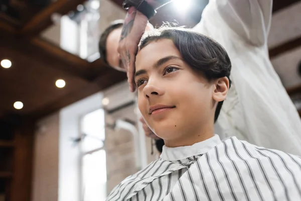 Low angle view of smiling teenage guy near blurred hairstylist in barbershop — Stock Photo