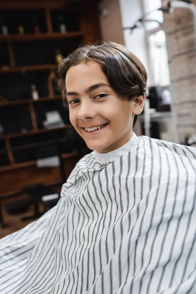 Felice adolescente in mantella parrucchiere guardando la fotocamera in barbiere — Foto stock