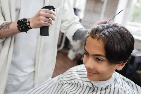 Татуированный парикмахер наносит лак для волос на улыбающегося клиента-подростка — стоковое фото
