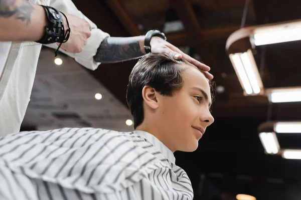 Vista de ángulo bajo de peluquero tatuado tocando el pelo de chico morena en la barbería - foto de stock