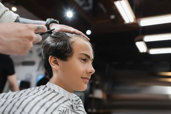 Vista lateral do menino adolescente na capa de cabeleireiro perto do barbeiro no salão de beleza — Fotografia de Stock