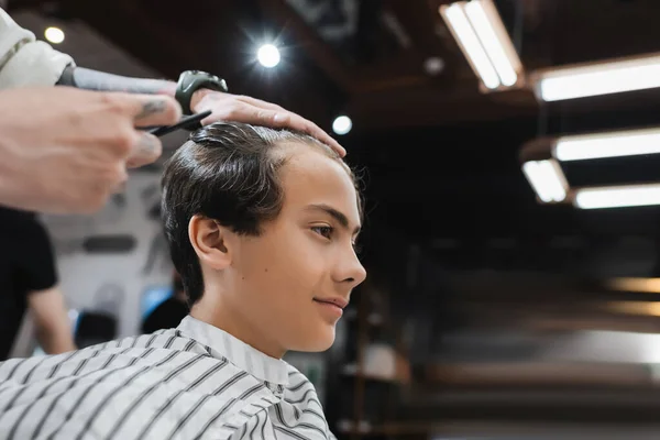 Vista lateral del adolescente en la capa de peluquería cerca de barbero en el salón de belleza - foto de stock