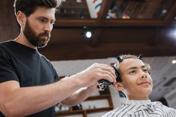 Бородатий перукар обрізає волосся позитивного підліткового клієнта в салоні краси — стокове фото