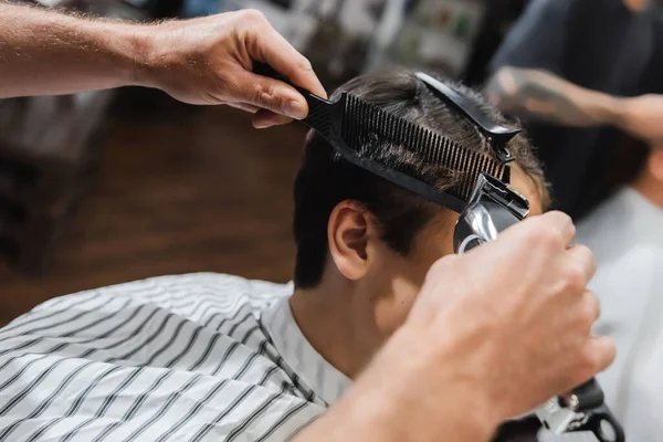 Hairdresser holding comb and clipper while trimming hair of teen boy in beauty salon — Stock Photo