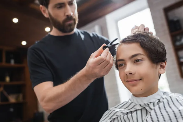 Sorrindo adolescente menino sentado perto de cabeleireiro borrado com grampo de cabelo no salão de beleza — Fotografia de Stock