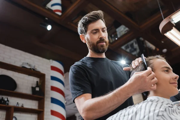 Vista de bajo ángulo de peluquería peinando el cabello de cliente adolescente en la barbería - foto de stock