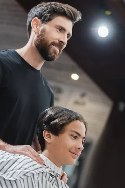 Улыбающийся парикмахер и подросток в плаще, смотрящий в салон красоты — стоковое фото