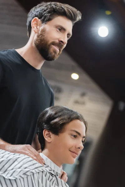 Coiffeur souriant et adolescent garçon en cape regardant loin dans le salon de beauté — Photo de stock