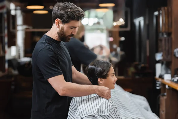 Vue latérale du coiffeur portant une cape de coiffure sur un client adolescent dans un salon de beauté — Photo de stock