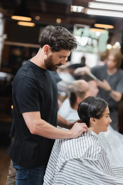 Vue latérale du coiffeur barbu portant une cape sur un client adolescent dans un salon de beauté — Photo de stock