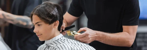 Cabeleireiro segurando cortador e pente perto adolescente na barbearia, banner — Fotografia de Stock