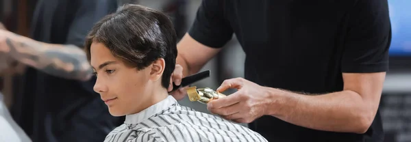 Peluquería sujetando clipper y peine cerca de adolescente en la barbería, pancarta - foto de stock