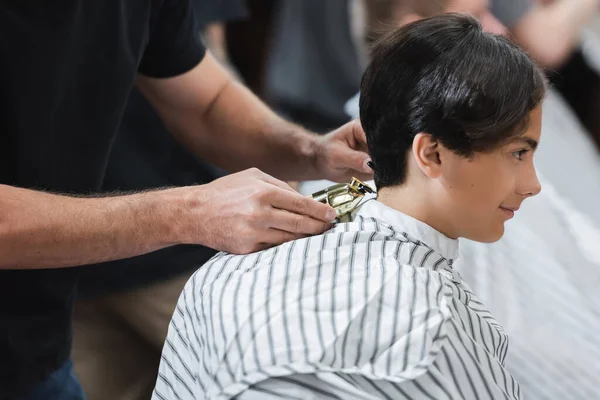 Парикмахер держит клиппер рядом с волосами подростка в плаще в салоне красоты — стоковое фото