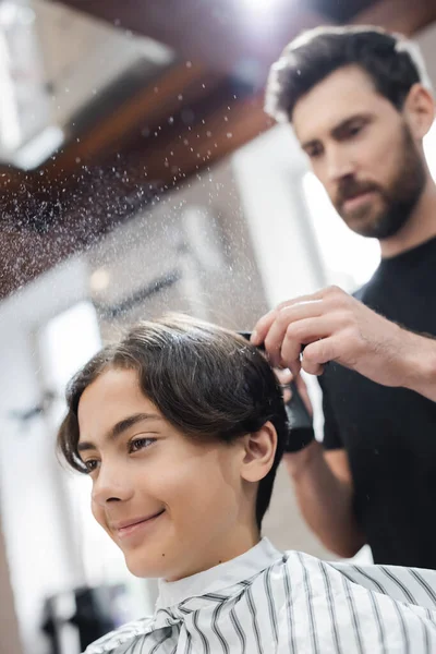 Cliente sorridente sentado perto de cabeleireiro borrado com pulverizador no salão de beleza — Fotografia de Stock