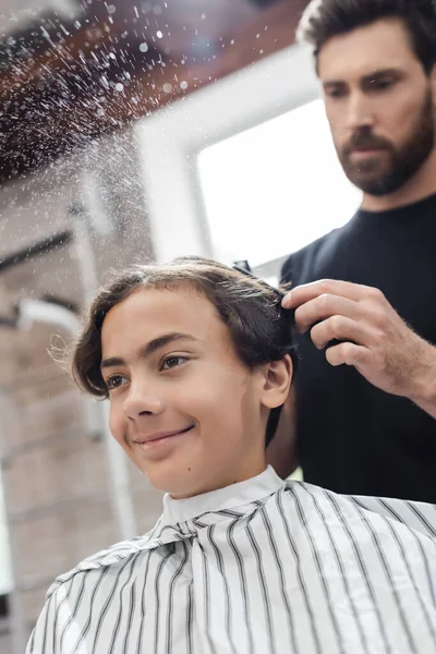 Adolescente alegre na capa sentado perto de cabeleireiro borrado pulverizando água no salão de beleza — Fotografia de Stock