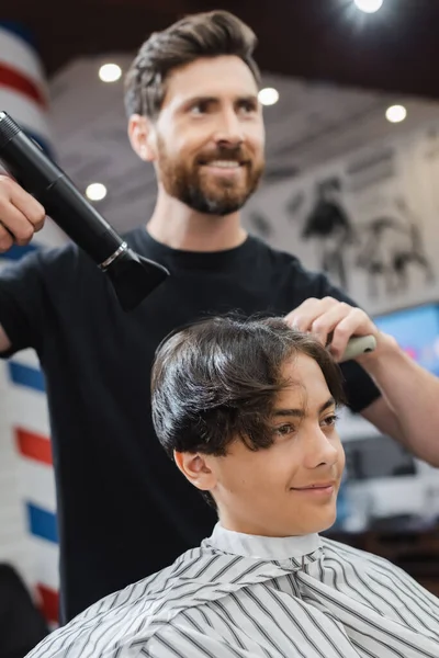 Cabeleireiro desfocado que seca o cabelo do rapaz adolescente na capa no salão de beleza — Fotografia de Stock