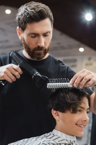 Barbeiro secagem e pentear o cabelo do menino adolescente sorridente no salão de beleza — Fotografia de Stock