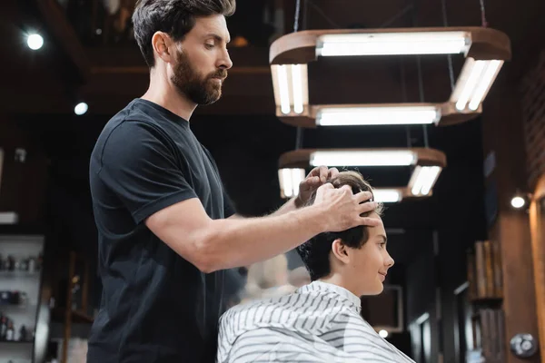 Vista lateral de peluquería peinado pelo de adolescente en capa en la barbería - foto de stock