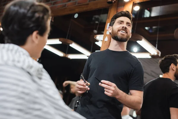 Low angle view of smiling hairstylist holding comb and scissors near blurred teen boy in beauty salon — Stock Photo