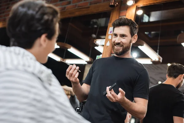 Smiling hairdresser holding scissors and comb near blurred teen boy in cape in beauty salon — Stock Photo