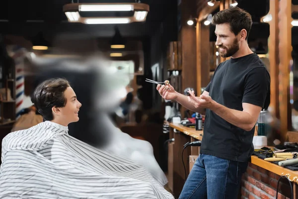 Positivo adolescente menino na capa olhando para cabeleireiro com tesoura e pente no salão de beleza — Fotografia de Stock
