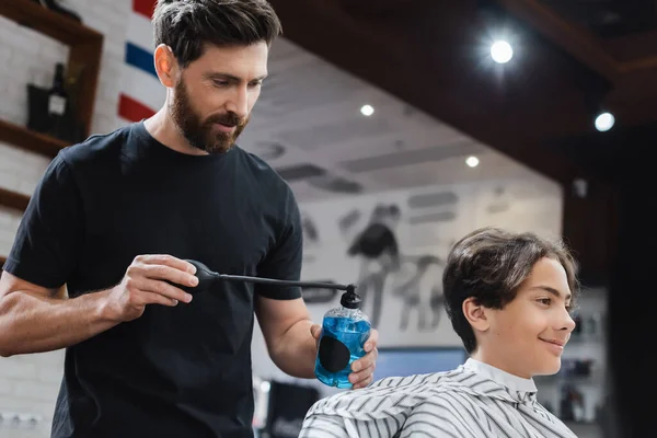 Parrucchiere barbuto utilizzando lacca per capelli sul ragazzo adolescente sorridente nel salone di bellezza — Foto stock