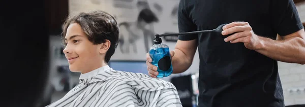 Hairstylist che tiene lacca per capelli vicino a ragazzo adolescente sorridente nel salone di bellezza, banner — Foto stock