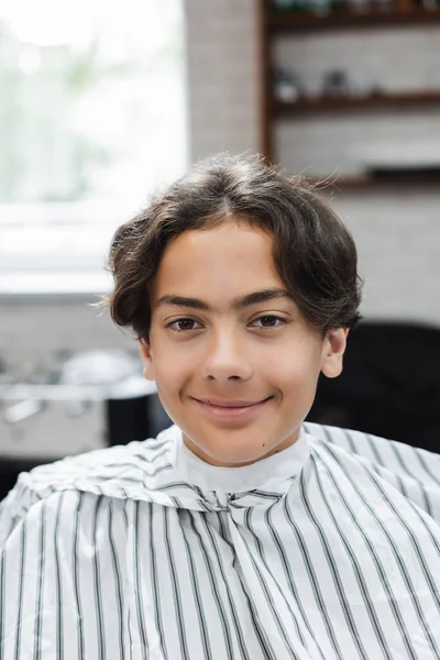 Smiling teenager in hairdressing cape looking at camera in beauty salon — Stock Photo