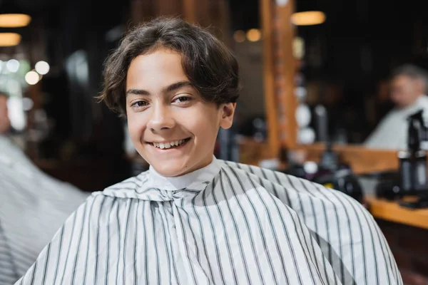 Cheerful teenager in hairdressing cape looking at camera in beauty salon — Stock Photo