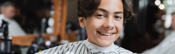 Positive teenager in hairdressing cape looking at camera in barbershop, banner — Stock Photo