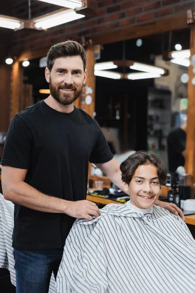 Barbeiro sorridente e adolescente olhando para a câmera na barbearia — Fotografia de Stock