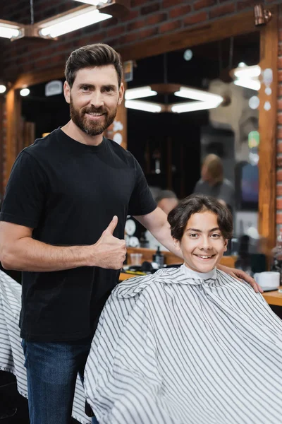 Позитивний перукар, що показує як жест і дивиться на камеру біля підлітка в перукарській накидці — стокове фото