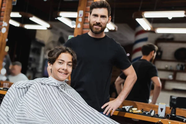 Smiling teenage boy in hairdressing cape looking at camera near bearded hairstylist in beauty salon — Stock Photo