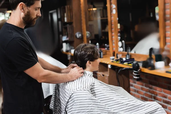 Side view of hairdresser wearing cape on teen boy in beauty salon — Stock Photo