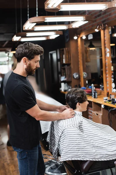 Vista lateral del peluquero con capa de peluquería en el adolescente en la barbería - foto de stock