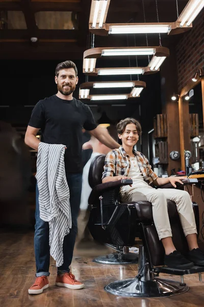 Coiffeur souriant tenant cap près de l'adolescent dans un fauteuil dans un salon de beauté — Photo de stock