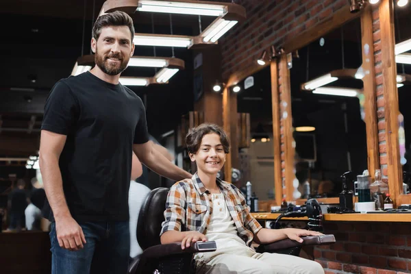 Позитивный парикмахер и подросток смотрит в камеру в парикмахерской — стоковое фото