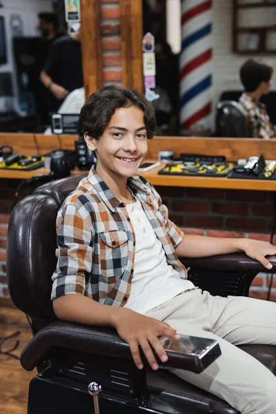 Happy teenage boy looking at camera while sitting on armchair in barbershop — Stock Photo