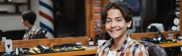 Adolescente sonriendo a la cámara mientras está sentado en el sillón en la barbería, pancarta - foto de stock