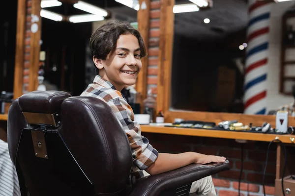 Sonriente adolescente mirando a la cámara mientras está sentado en el sillón en el salón de belleza - foto de stock