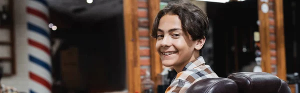 Positivo adolescente menino sorrindo para a câmera enquanto sentado em poltrona no salão de beleza, banner — Fotografia de Stock